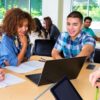 Diverse group of male and female teens and young adults are studying together around desks in classroom or library. Caucasian, African American and Hispanic students are looking at laptop computer screen and working on assignment together.