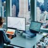 Back view of business woman sitting at panoramic skyscraper office desktop front PC computer with financial graphs and statistics on monitor.Analysis of digital market and investment in block chain